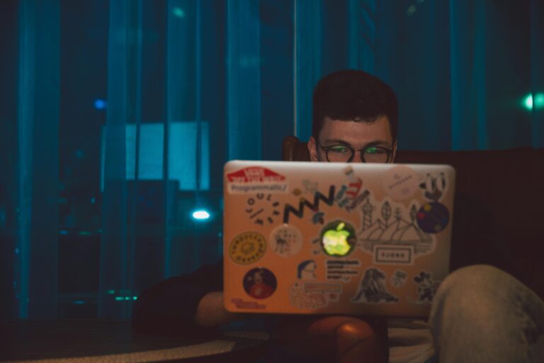 A man focused on working on his laptop with stickers at night, indoors.